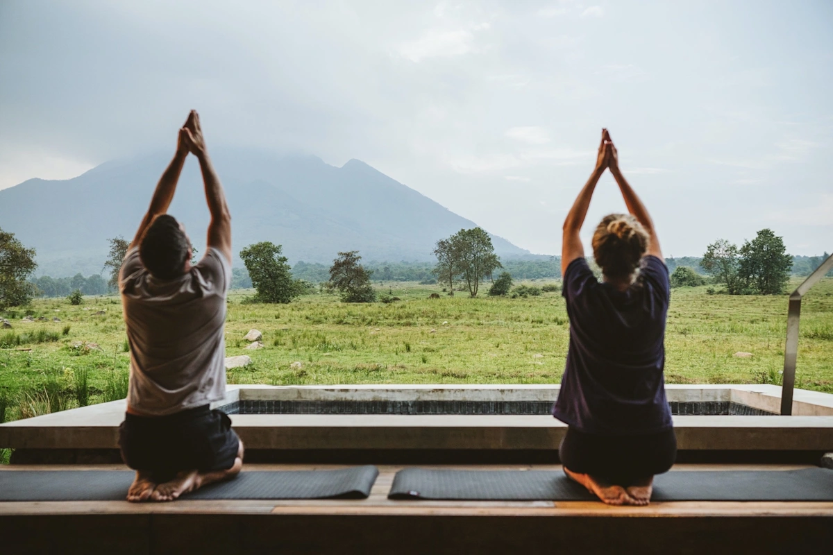 A guest enjoys a wellness activity at Kwitonda Lodge, surrounded by lush greenery and tranquil natural surroundings, with the mist-covered volcanic peaks of Rwanda’s Volcanoes National Park visible in the background. The serene setting combines luxury, relaxation, and breathtaking views of the park’s iconic mountain landscape.